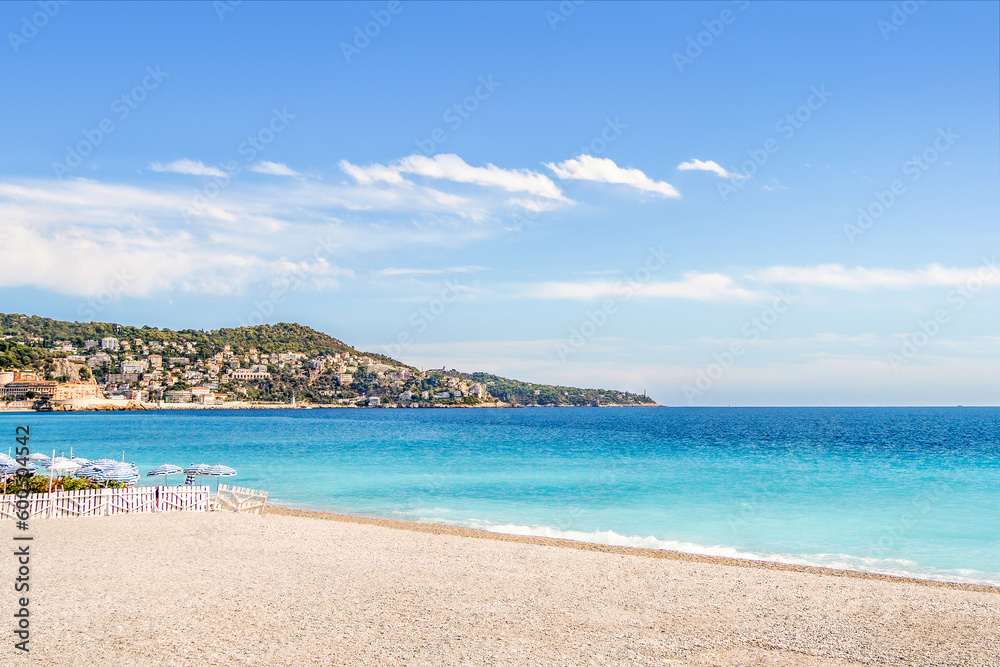 Beach, sea and coast in Nice, South of France. French Riviera shore. Blue sky and turquoise water. Mountain village and town in the background. Bright summer landscape of mediterranean destination.