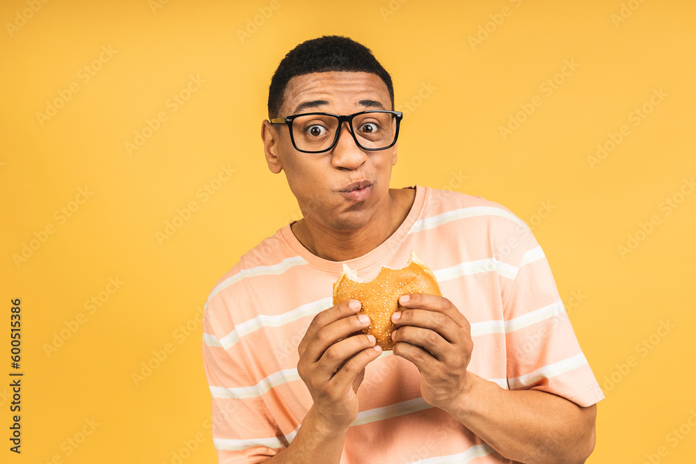 Hungry young african american man eating hamburger or burger isolated over yellow background. Diet concept. Hungry young african american man eating hamburger or burger isolated over yellow background. Diet concept.