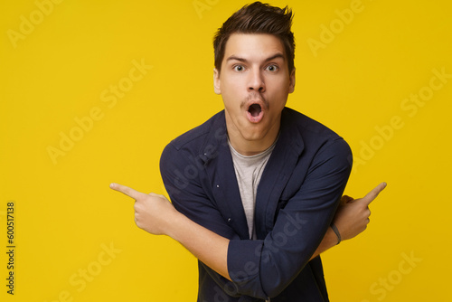 Surprised young man is depicted pointing with two crossed hands in opposite directions against yellow background, with plenty of copy space for text. Sense of astonishment, amazement, or shock, with photo