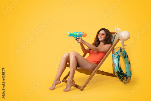 Glad millennial asian lady in swimsuit, sunglasses sit on deck chair, enjoying free time and playing water gun