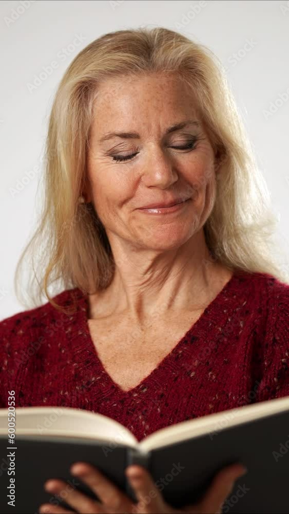 Vertical video reading then looking into camera, happy pretty elderly blond woman 50s years old wears sweater reading book isolated model on solid white background studio portrait with copy space Vertical video reading then looking into camera, happy pretty elderly blond woman 50s years old wears sweater reading book isolated model on solid white background studio portrait with copy space