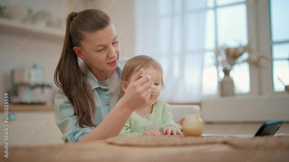 Mother feeds her baby fruit or vegetable puree using plastic spoon. Toddler eats with pleasure. Mom holds child on knees at table on kitchen. Raising child, kid's nutrition and motherhood concept. Mother feeds her baby fruit or vegetable puree using plastic spoon. Toddler eats with pleasure. Mom holds child on knees at table on kitchen. Raising child, kid's nutrition and motherhood concept.