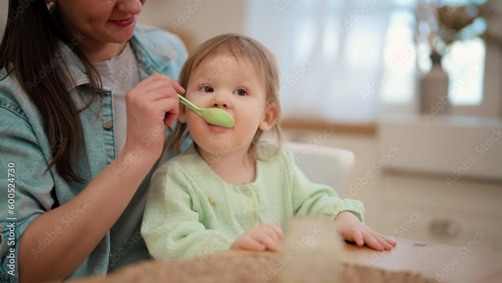 Mother feeds her baby toddler dairy food yogurt from plastic container on kitchen at home sitting at table. Mom holds her little kid with dirty face on knees. Raising child, kid's nutrition concept. Mother feeds her baby toddler dairy food yogurt from plastic container on kitchen at home sitting at table. Mom holds her little kid with dirty face on knees. Raising child, kid's nutrition concept.