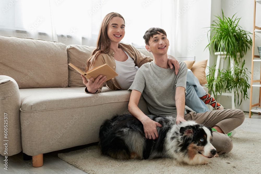 cheerful gay man sitting on carpet and cuddling Australian shepherd dog next to happy partner with long hair holding book and looking at camera in modern living room cheerful gay man sitting on carpet and cuddling Australian shepherd dog next to happy partner with long hair holding book and looking at camera in modern living room
