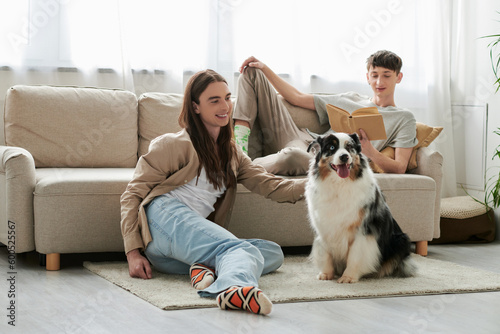 happy gay man reading book and lying on comfortable couch while his boyfriend with long hair playing with Australian shepherd dog during his free time at home