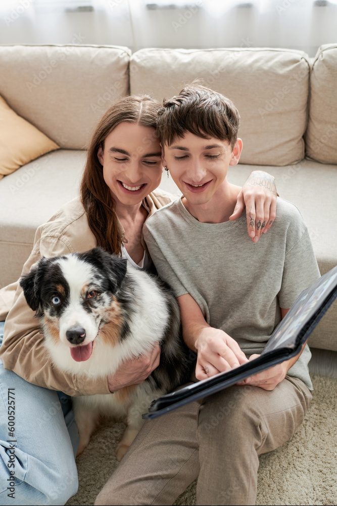 happy gay couple looking together at photo album and smiling while having happy memories and sitting near Australian shepherd dog and sofa in modern living room