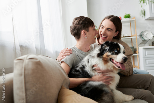 Young overjoyed same sex couple in casual clothes talking and hugging near furry Australian shepherd dog while resting on couch in living room at home © LIGHTFIELD STUDIOS
