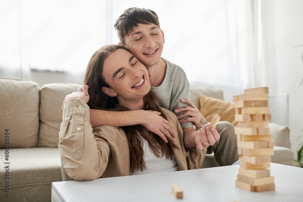 Young smiling gay man with closed eyes hugging long haired boyfriend in casual clothes near blurred wood blocks game on table in living room at home Young smiling gay man with closed eyes hugging long haired boyfriend in casual clothes near blurred wood blocks game on table in living room at home