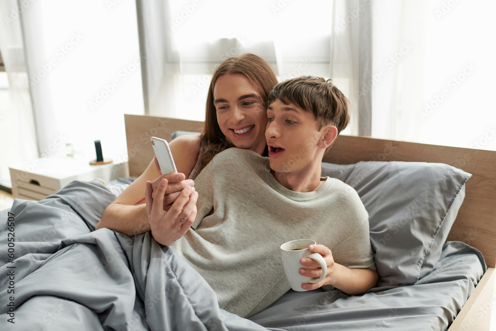 Smiling long haired gay man showing smartphone at excited boyfriend in pajama holding coffee cup while lying under blanket on bed at home in morning Smiling long haired gay man showing smartphone at excited boyfriend in pajama holding coffee cup while lying under blanket on bed at home in morning