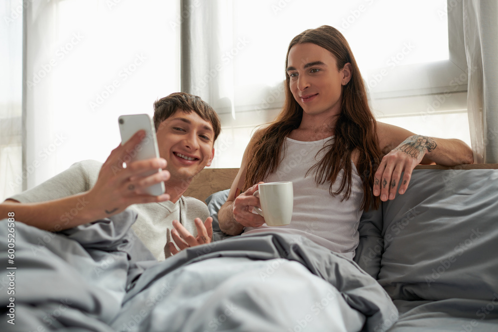 Carefree gay man in pajama holding mobile phone near long haired and tattooed boyfriend holding coffee cup and sitting on bed at home in morning Carefree gay man in pajama holding mobile phone near long haired and tattooed boyfriend holding coffee cup and sitting on bed at home in morning