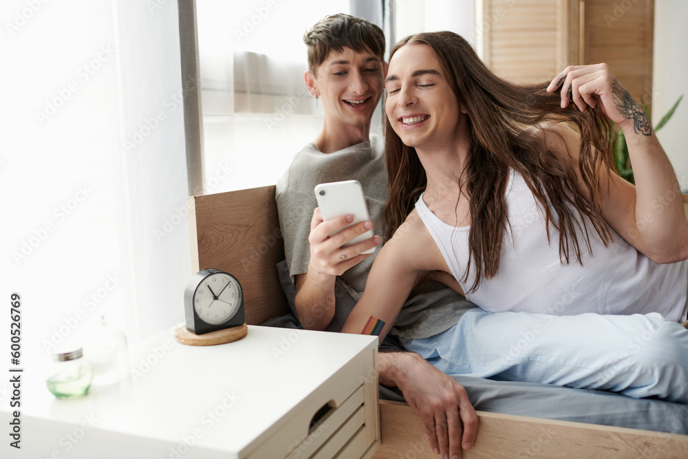 Smiling lgbt couple in sleepwear using mobile phone together while relaxing on cozy bed near alarm clock on bedside table in morning at home Smiling lgbt couple in sleepwear using mobile phone together while relaxing on cozy bed near alarm clock on bedside table in morning at home
