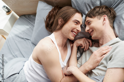 Top view of cheerful and tattooed homosexual couple in sleepwear talking with each other while lying together on bed in modern bedroom in morning at home