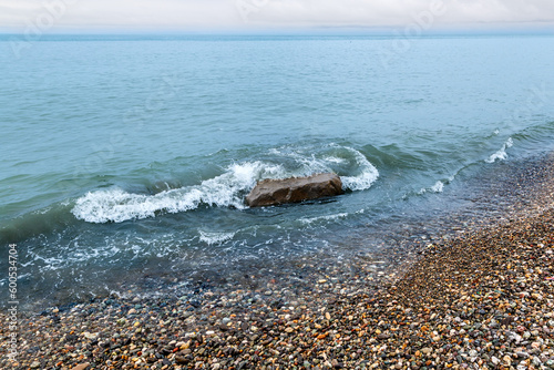 Emerald-green sea waves wash over a large stone lying on a pebble beach
