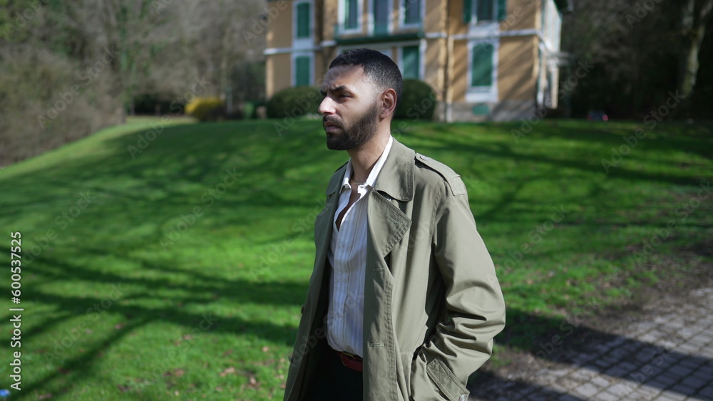 One confident young Middle Eastern man walking outside at park wearing fashionable jacket. An Arab male person with serious expression walks outdoors