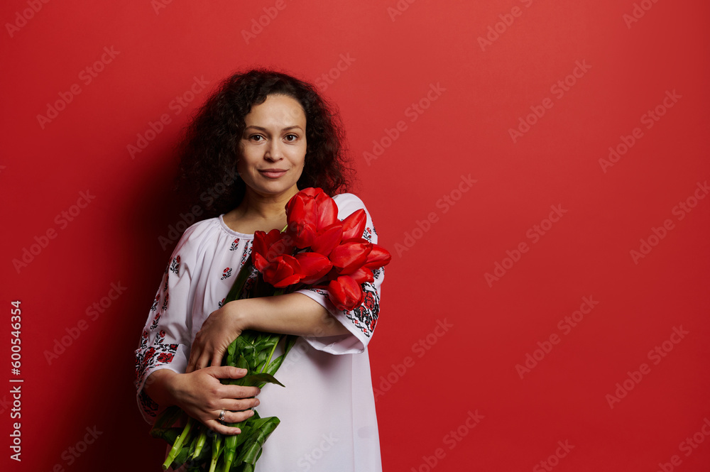 Beautiful woman in embroidered white dress, holds a bouquet of red tulips, smiles looking at camera, isolated on red background. Copy space. Ukrainian style, culture and traditions. Independence Day Beautiful woman in embroidered white dress, holds a bouquet of red tulips, smiles looking at camera, isolated on red background. Copy space. Ukrainian style, culture and traditions. Independence Day