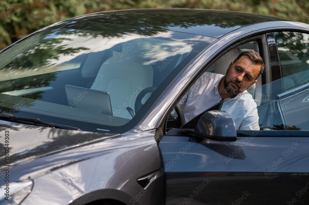 Caucasian bearded man in a suit gets out of a black electro car in the countryside in summer.