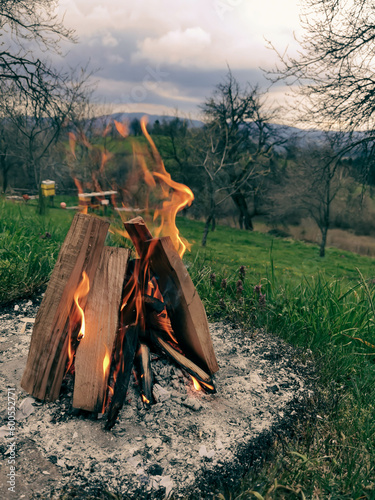 Big bonfire fire on wild nature background with stones. Close-up travel, adventure hot and bright flame light. Cozy evening in the forest outdoors. Burning wooden firewood, log, timber.