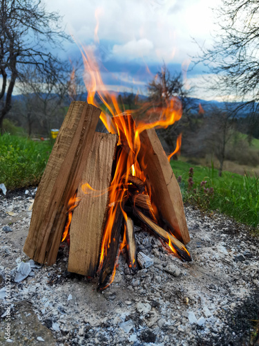 Big bonfire fire on wild nature background with stones. Close-up travel, adventure hot and bright flame light. Cozy evening in the forest outdoors. Burning wooden firewood, log, timber.