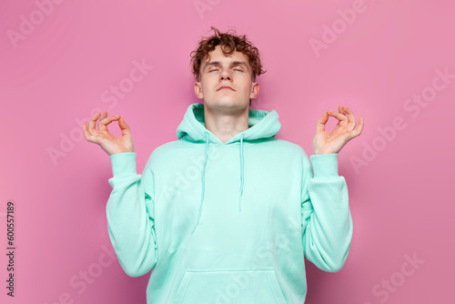 young curly guy in mint hoodie meditates in lotus position on pink isolated background, calm man in nirvana