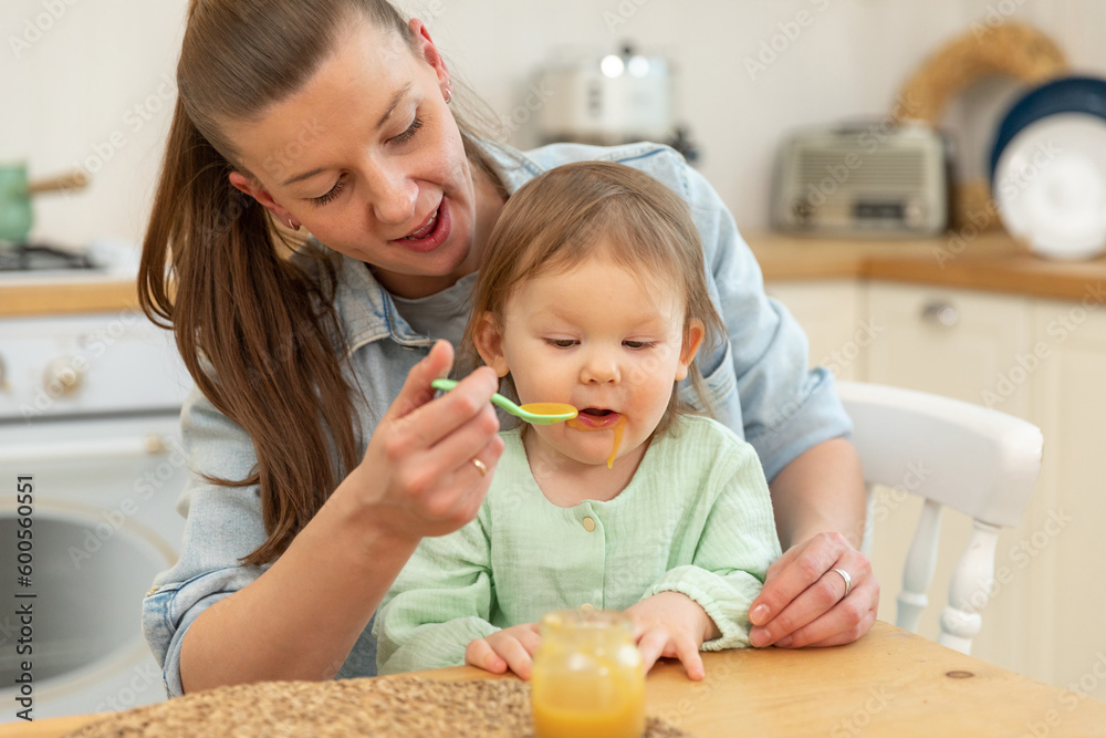 Happy family at home. Mother feeding her baby girl from spoon in kitchen. Little toddler child with messy funny face eats healthy food at home. Young woman mom giving food to kid daughter Happy family at home. Mother feeding her baby girl from spoon in kitchen. Little toddler child with messy funny face eats healthy food at home. Young woman mom giving food to kid daughter
