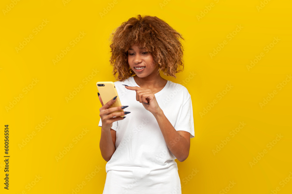 cute curly american girl with braces in white t-shirt uses smartphone and smiles on yellow isolated background cute curly american girl with braces in white t-shirt uses smartphone and smiles on yellow isolated background