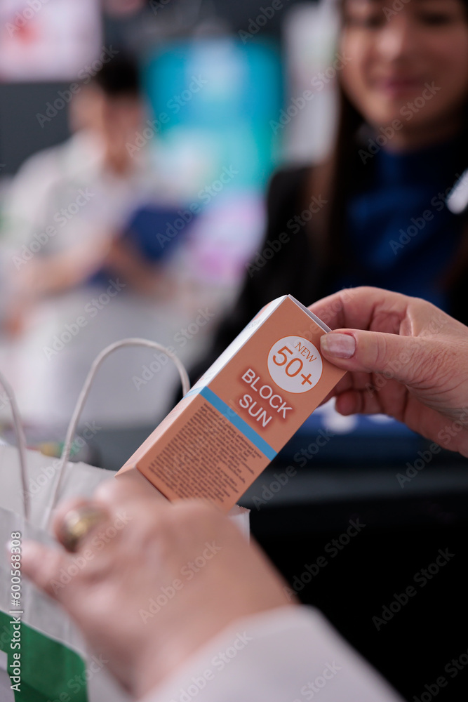 Pharmacy cashier selling sunblock lotion and putting product in buyer shopping bag. Medical retail store seller holding sunscreen, throwing cosmetology sun protection cream in paper package close up