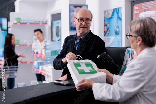 Senior man taking medications order from pharmacist at checkout. Elderly client buying supplements and disease remedy in pharmacy store, consulting with drugstore employee at cash register
