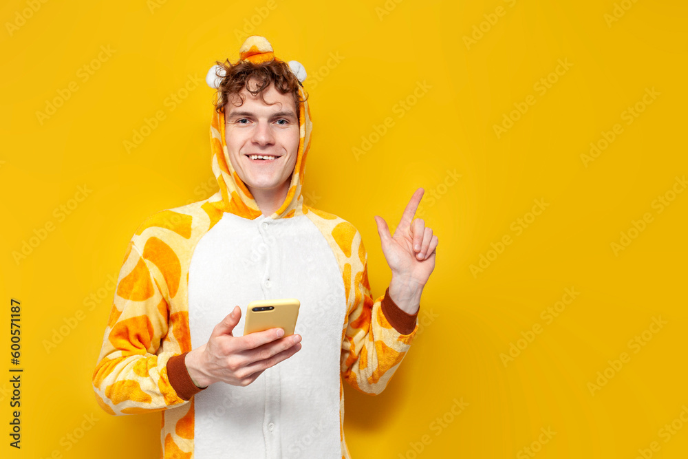 young curly guy in funny giraffe pajamas uses smartphone and shows his hand to the side on yellow background young curly guy in funny giraffe pajamas uses smartphone and shows his hand to the side on yellow background