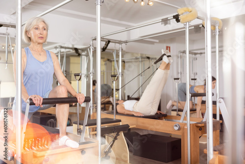 Positive senior woman doing stretching exercises on pilates reformer as part of injury rehabilitation course. Therapeutic physical training concept