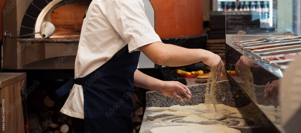 Male chef kneading and rotating dough basis for pizza on the grey marble cooking table in pizzeria's kitchen table