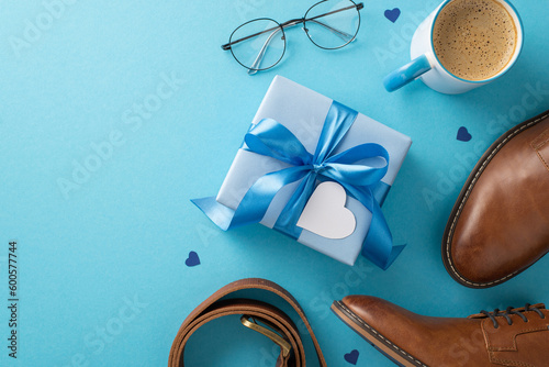 Chic Father's Day theme. Overhead shot of present box, dress shoes, accessories, belt, glasses, coffee cup on blue backdrop with blank space for text