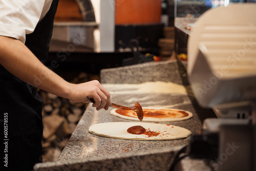 Anonymous male chef spreading tomato sauce with big iron spoon onto pizza while preparing pizza in pizzeria kitchen. High quality photo