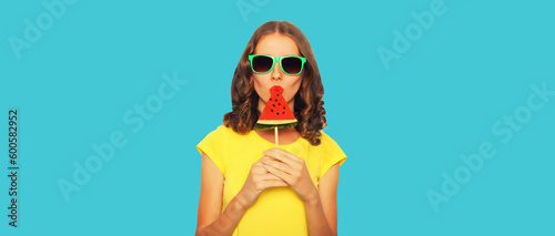 Summer portrait of happy young woman with fresh juicy fruits, girl eating lollipop or ice cream shaped slice of watermelon on blue background
