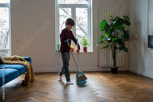 Teen boy doing chores cleaning floor in living room sweeping trash with broom to scoop. Teenager helping with household duties. Tidying up house involving children in family to maintain order at home.