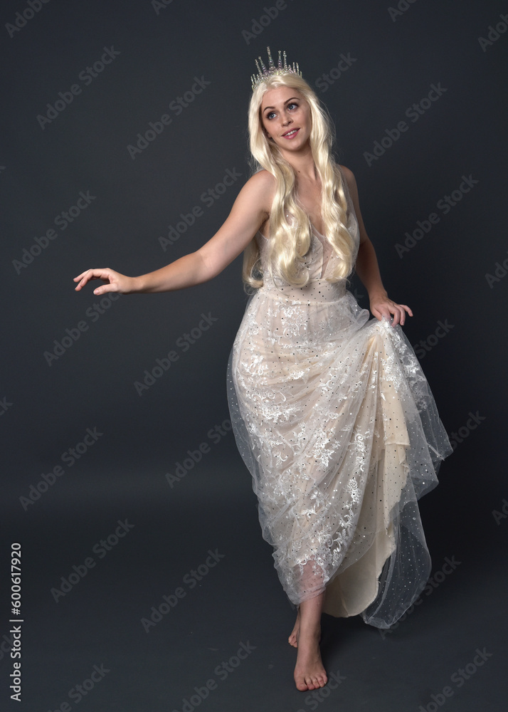 Fototapeta premium Full length portrait of beautiful women with long blonde hair, wearing fantasy princess crown and elegant white ball gown, standing pose with hand gesture. Isolated on dark grey studio background.