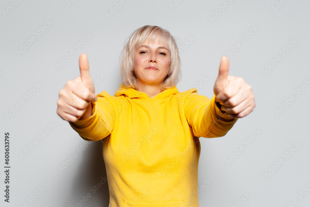 Studio portrait of happy blonde woman showing thumbs up on white background, wearing yellow sweater. Studio portrait of happy blonde woman showing thumbs up on white background, wearing yellow sweater.