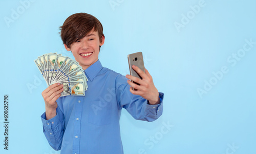 A young smiling teenage man in a blue shirt takes a selfie on a mobile phone, holding a fan of cash money in dollar banknotes, isolated on a blue background, copy space. People lifestyle concept.