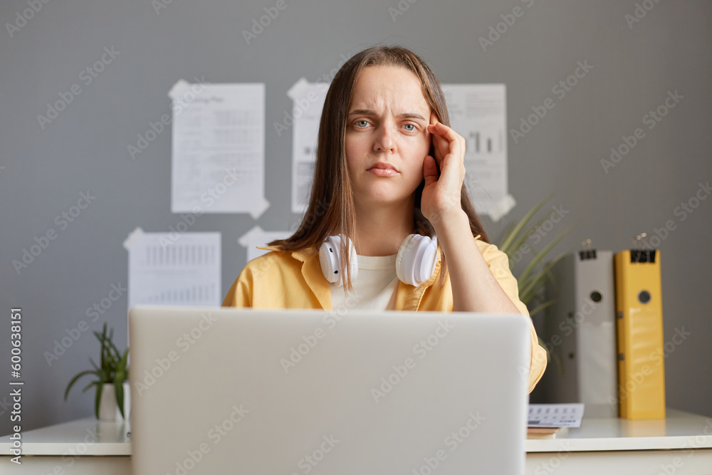 Tired woman sitting in front of computer feeling fatigue suffering headache spending long hours doing online work holding laptop massaging temple looking at camera. Tired woman sitting in front of computer feeling fatigue suffering headache spending long hours doing online work holding laptop massaging temple looking at camera.