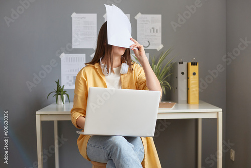 Tired exhausted woman working with documents and laptop having job troubles covering face with papers making mistake expressing negative feelings. photo