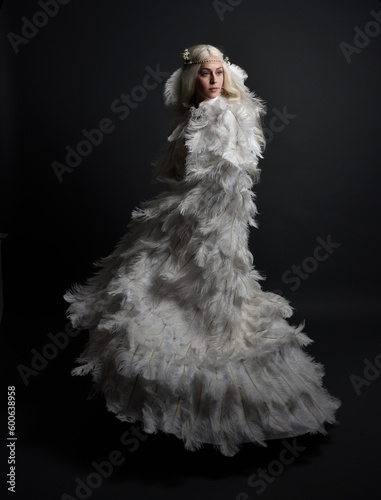 fantasy portrait of beautiful female model with long blond hair wearing otherworldly  white feathered cloak costume and headdress, isolated on dark studio background.