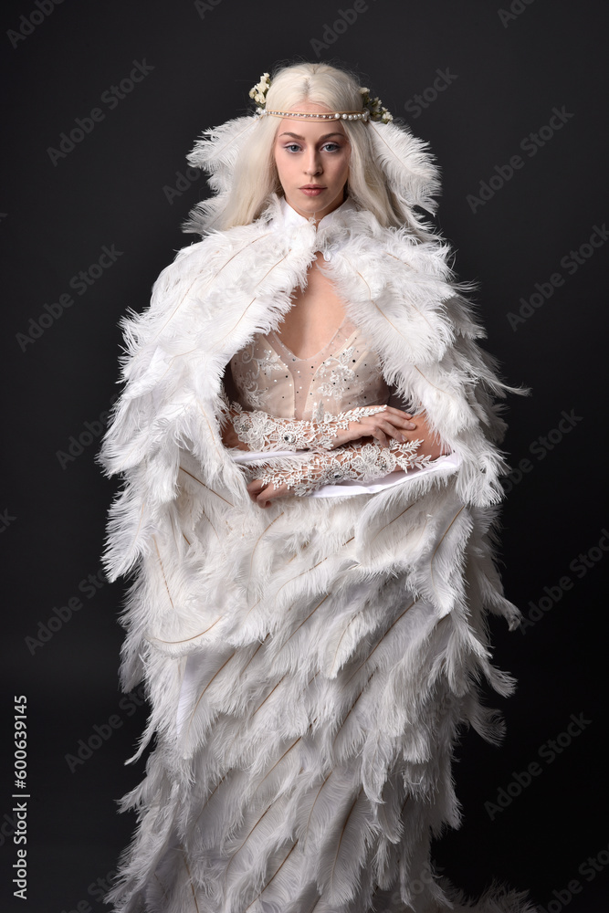 fantasy portrait of beautiful female model with long blond hair wearing otherworldly white feathered cloak costume and headdress, isolated on dark studio background. fantasy portrait of beautiful female model with long blond hair wearing otherworldly white feathered cloak costume and headdress, isolated on dark studio background.