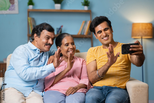 Happy smiling indian senior parents with adult son making video call on mobile phone while sitting on sofa at home - concept of technology, family bonding and distant connection.