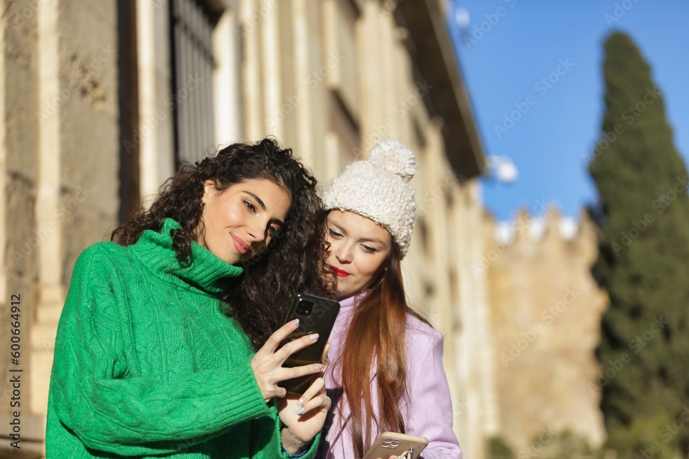 Two women, young and beautiful, with sweater, coat and one with wool hat, consulting social networks on their mobiles. Concept apps, smartphone, friendship, couples. Selective focus on the brown woman Two women, young and beautiful, with sweater, coat and one with wool hat, consulting social networks on their mobiles. Concept apps, smartphone, friendship, couples. Selective focus on the brown woman