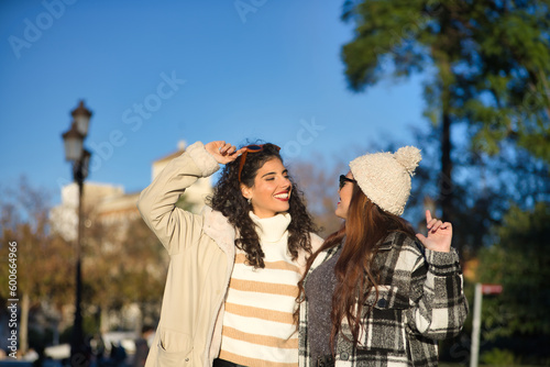 Two women, young and beautiful, with sweater, sunglasses, coat and one with wool hat, embraced, looking at each other, very smiling and happy. Concept autumn, winter, cold, friendship, full happiness.