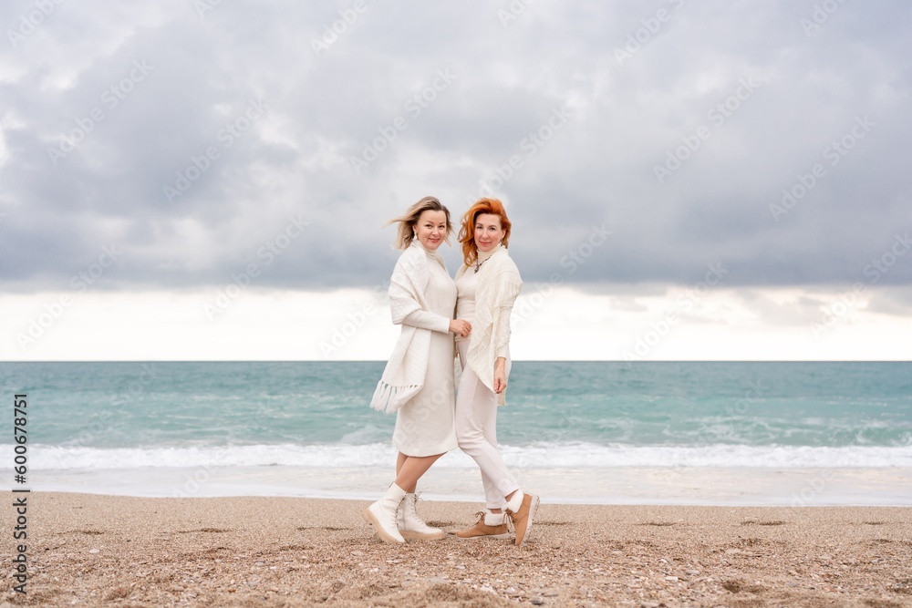 Women sea walk friendship spring. Two girlfriends, redhead and blonde, middle-aged walk along the sandy beach of the sea, dressed in white clothes. Against the backdrop of a cloudy sky and the winter