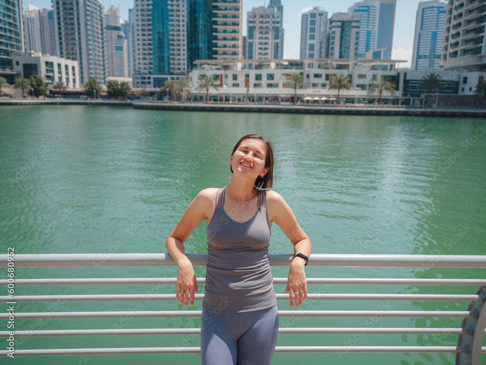 Beautiful young woman standing and Dubai marina skyline. having rest after morning run at the city. Portrait of satisfied woman after fitness exercises.