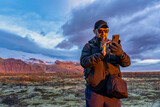 man holds a glove with teeth and using a phone for navigation with mountains with snow on background during sunset in iceland
