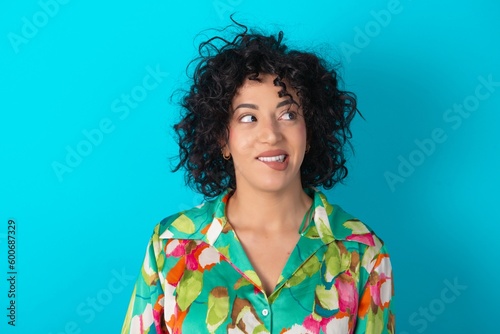 Amazed Young arab woman wearing colorful shirt over blue background bitting lip and looking tricky to empty space. © Roquillo