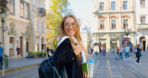 Pretty smiling happy modern young blond woman in casual clothes walking in the city center with ancient buildings while going to the work or study as she carrying textbooks in hands © serg