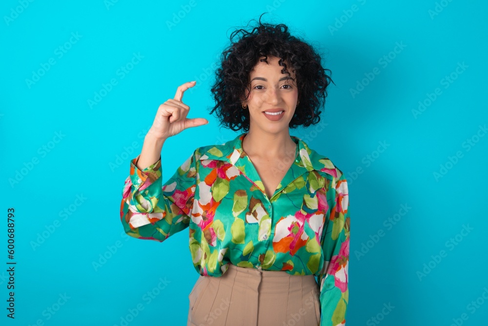 young arab woman wearing colorful shirt over blue background smiling and confident gesturing with hand doing small size sign with fingers looking and the camera. Measure concept young arab woman wearing colorful shirt over blue background smiling and confident gesturing with hand doing small size sign with fingers looking and the camera. Measure concept